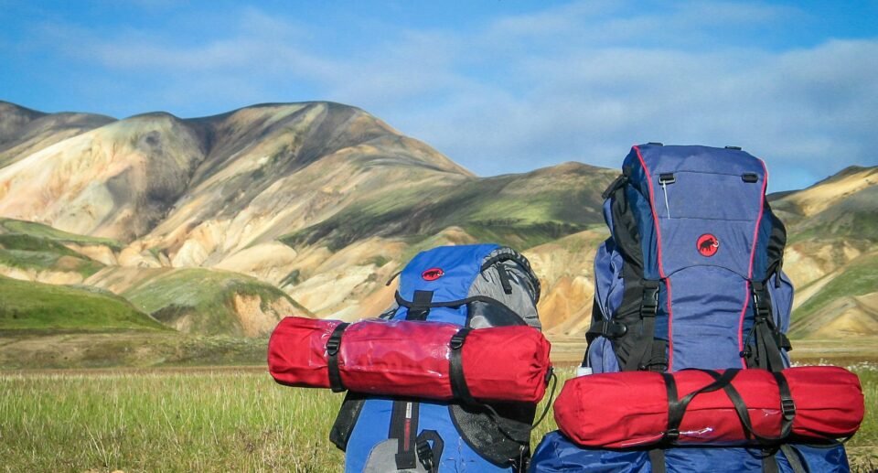 Two fully packed hiking backpacks with rolled sleeping mats resting in a grassy valley, set against colorful volcanic mountains under a clear blue sky, ready for a multi-day trekking adventure.