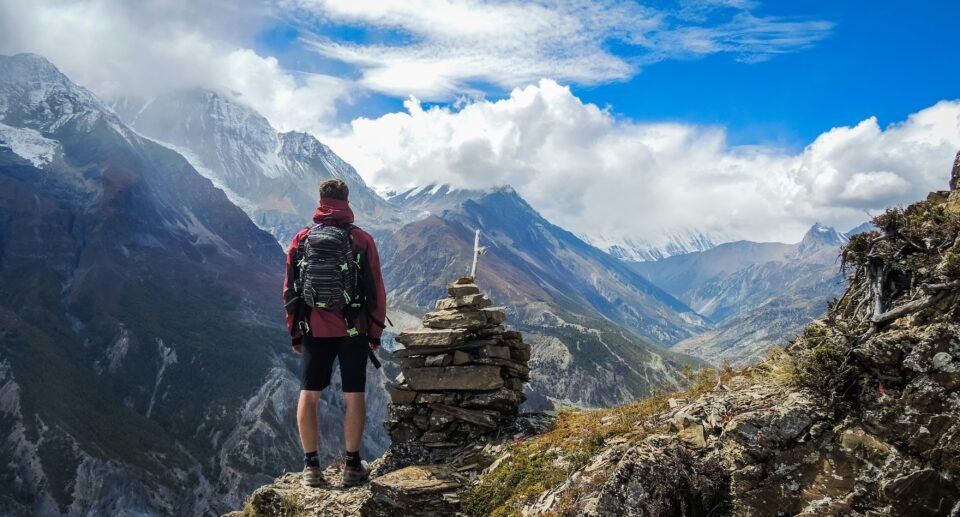 **Alt Text:** A lone backpacker standing on a rocky mountain viewpoint overlooking a dramatic alpine valley with snow-capped peaks, rugged cliffs, and billowing clouds under a bright blue sky, capturing the spirit of a multi-day trekking adventure.