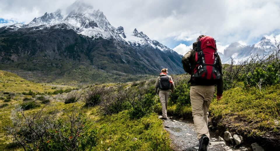 **Alt Text:** Two backpackers hiking along a mountain trail through a lush valley toward dramatic snow-capped peaks under a cloudy sky, capturing the spirit of a challenging multi-day trekking adventure in a remote alpine landscape.
